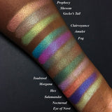 Top angled arm swatches on deep skin tone of Witchcraft vs. Alchemy Collection including Shroom Duochrome Eyeshadow next to Prophecy, Fog, Gecko's Tail, Clairvoyance, Amulet, Toadstool, Morgana, Hex, Salamander, Nocturnal, Eye of Newt