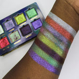 Arm swatches on deep skin tone of all 8 shadows included in the Deep Sea Treasures Palette with the Palette in the background. Top to bottom: Saltwater Pearl, The Bends, Fool's Gold, Kelp Forest, Ring of Fire, Cephalopod, Shipwreck and S.C.U.B.A.